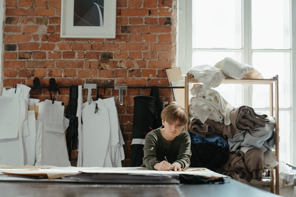 pexels-photo-4621565 A focused fashion designer draws patterns in a cozy, brick-walled atelier filled with fabrics.